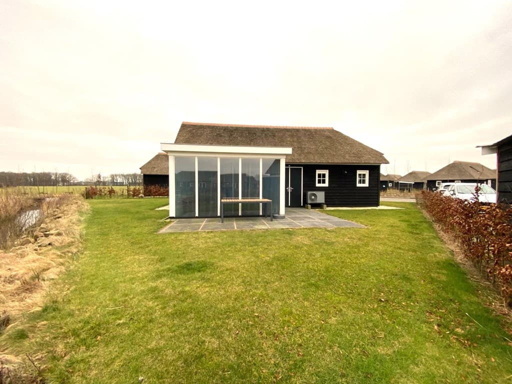 Zwarte vakantiewoning met rieten dak en terras met zithoek, omringd door gras en een haag.