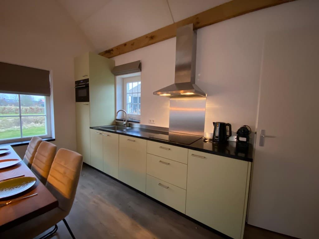 Keuken met lichte kasten, werkblad van zwart graniet, kookplaat, afzuigkap en koffiezetapparaat, achterraam met uitzicht op g
