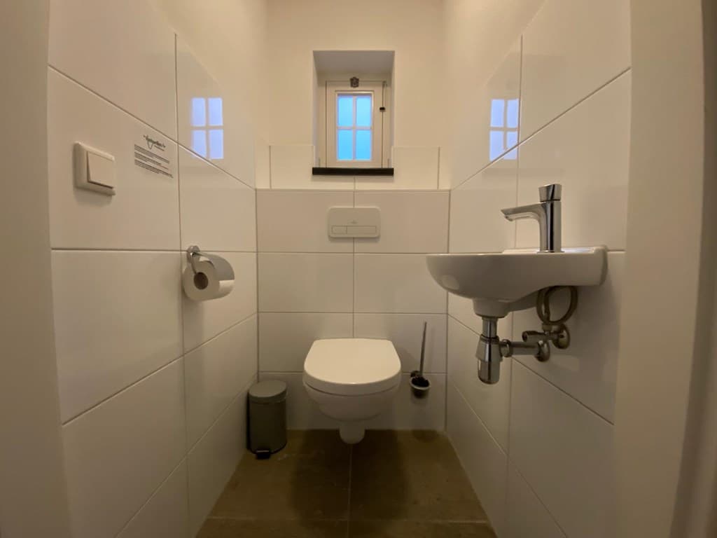 Witte betegelde toiletruimte met een toilet, wastafel en klein raam voor natuurlijk licht