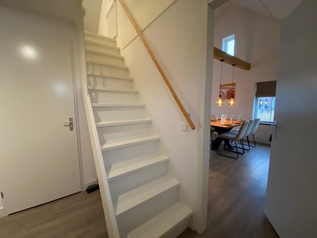 Wit trappenhuis met houten leuning en zicht op eetkamer met een lange tafel en hanglampen.