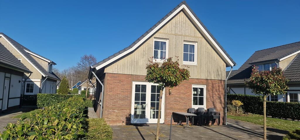 Modern huis met een houten gevel, terras met tuinmeubilair en omliggende groene hagen onder een blauwe lucht