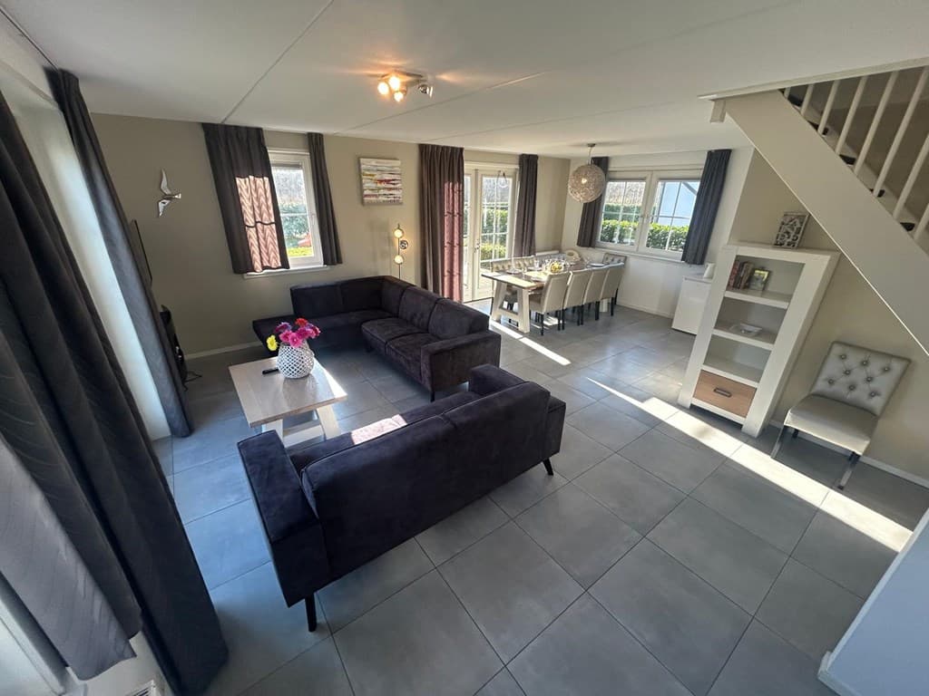 Moderne woonkamer met loungebanken, eettafel, grote ramen en trap naar de bovenverdieping in EuroParcs Limburg.