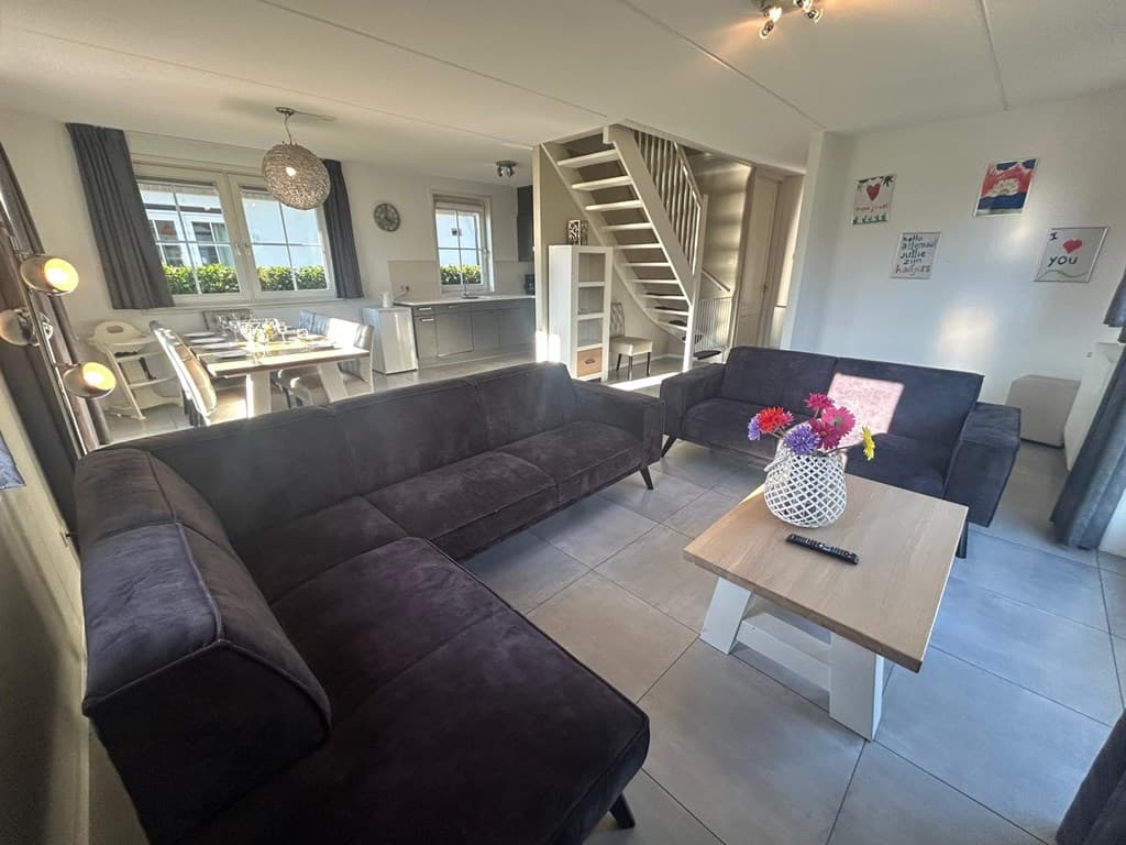 Zithoek met een donkere bank, salontafel en decoratieve bloemen in een vaas, toegang tot de keuken en eetruimte zichtbaar