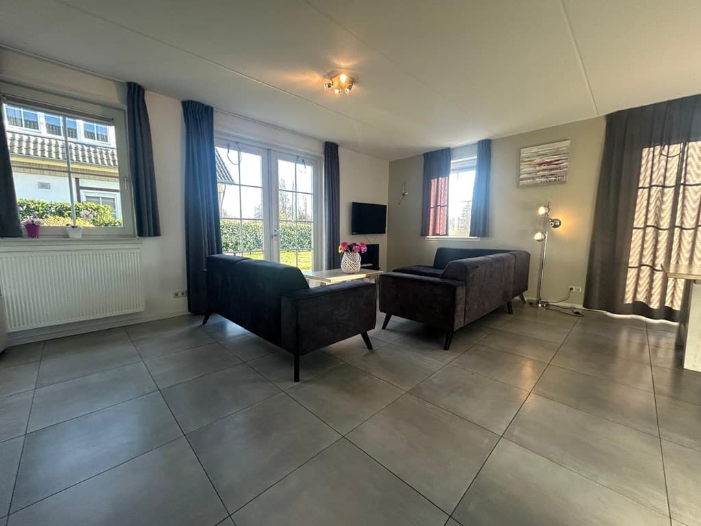 Woonkamer met twee banken, televisie aan de muur, vloertegels en grote ramen met uitzicht op de tuin