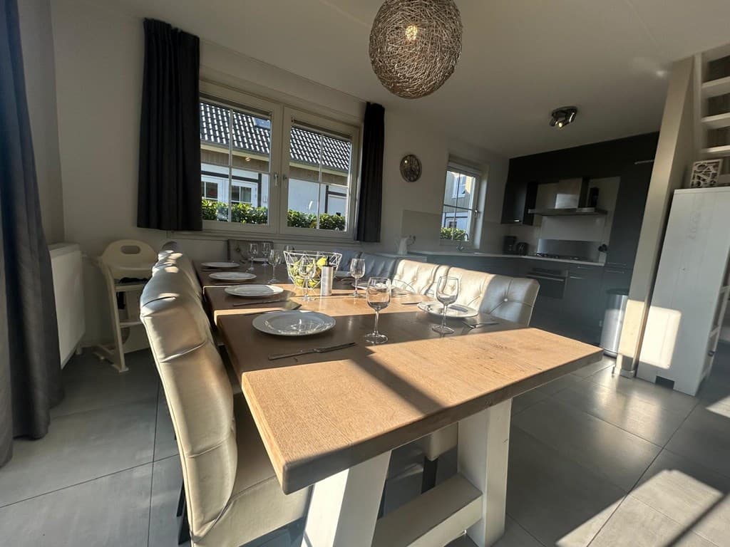 Eethoek met groot houten tafel, zorgvuldig dekken, en moderne keuken op de achtergrond met raam en gordijnen.