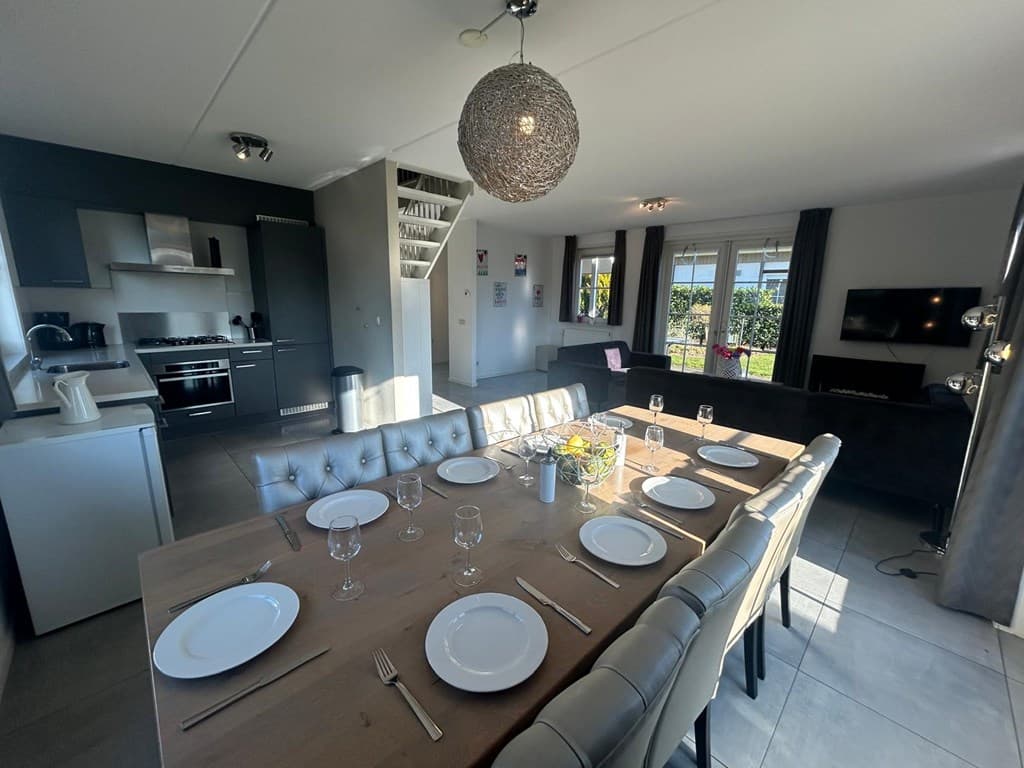 Eethoek met grote tafel en stoelen, moderne open keuken en zithoek met bank en tv in lichte ruimte met ramen.