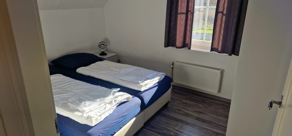 Slaapkamer met twee eenpersoonsbedden, nachtkastje, lamp en raam met gordijnen zichtbaar.