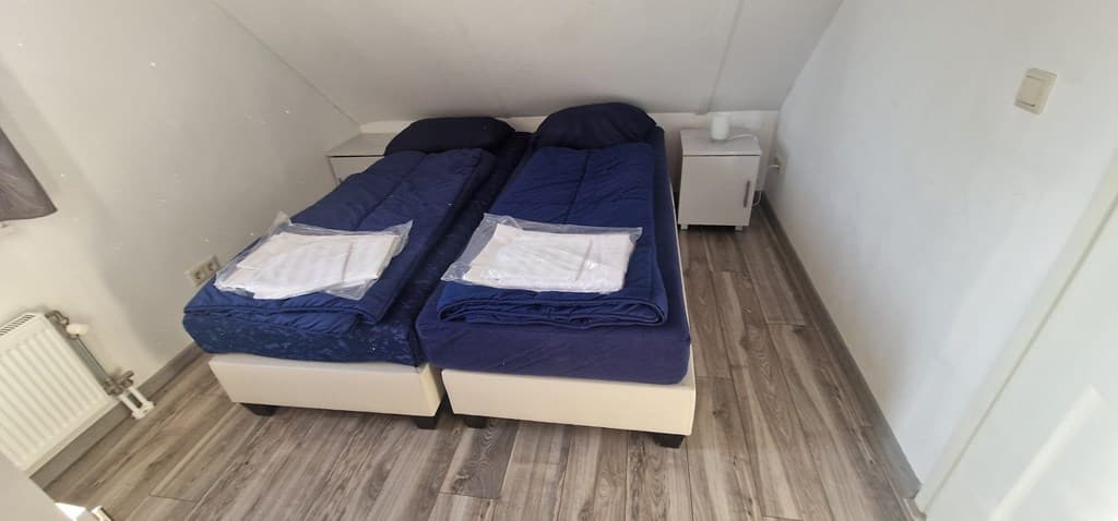 Twee eenpersoonsbedden met blauwe dekbedden in een lichte slaapkamer met laminaatvloer en nachtkastje.
