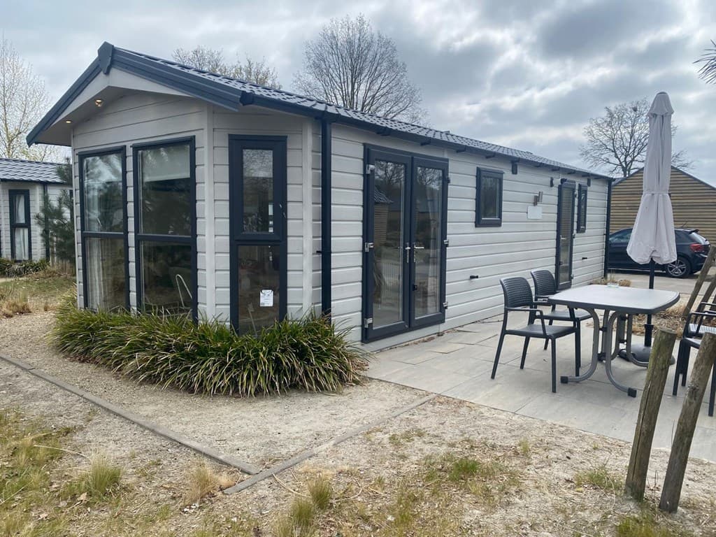 Moderne vakantiewoning met grote ramen en een terras, omgeven door groen en zandgrond.