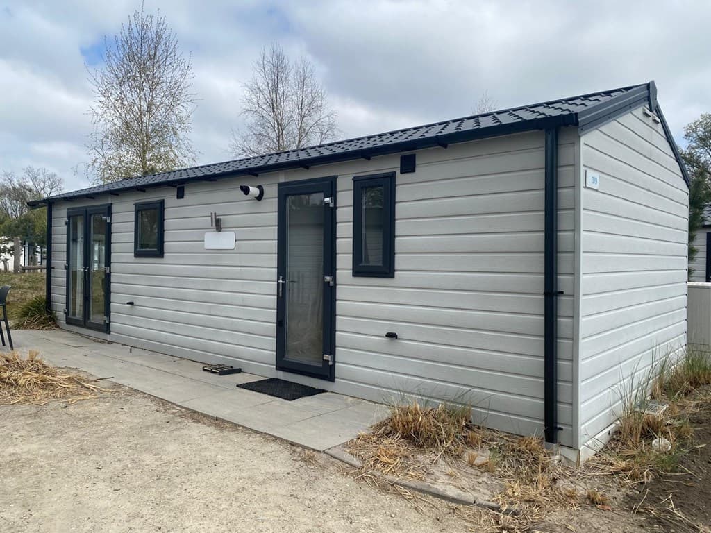 Een moderne vakantiewoning met een lichte gevel en een zwarte metalen dak, gelegen in een groene omgeving.