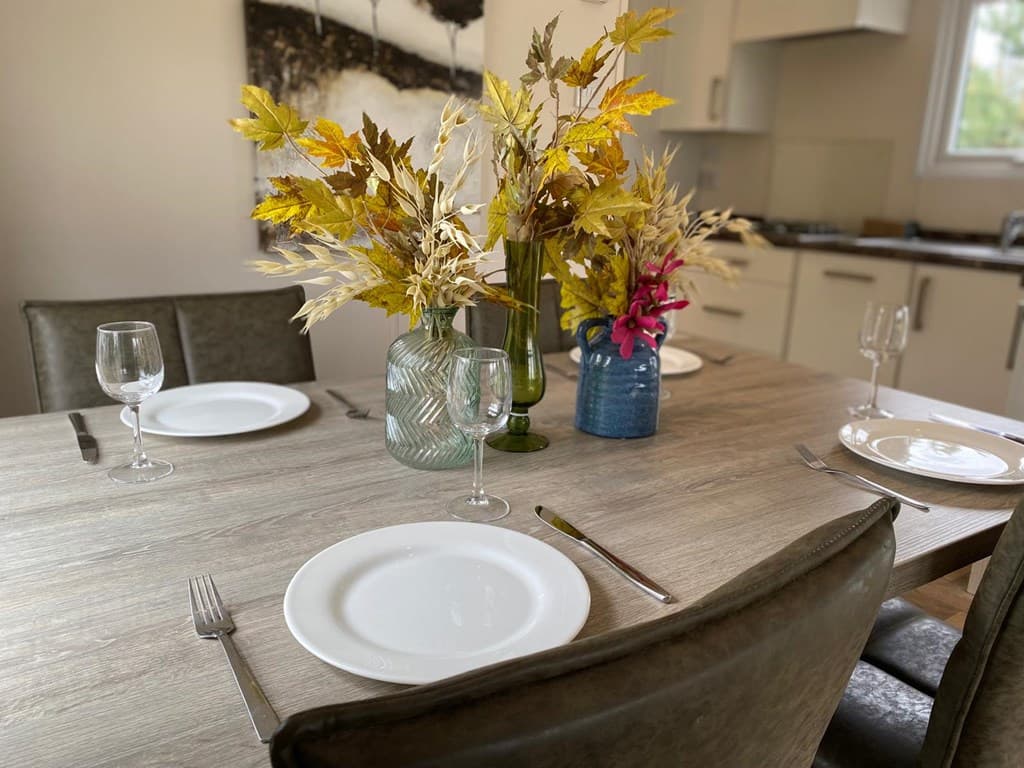 Eettafel met vier borden, glazen en vazen met droogbloemen in een lichte ruimte met een open keukentje.