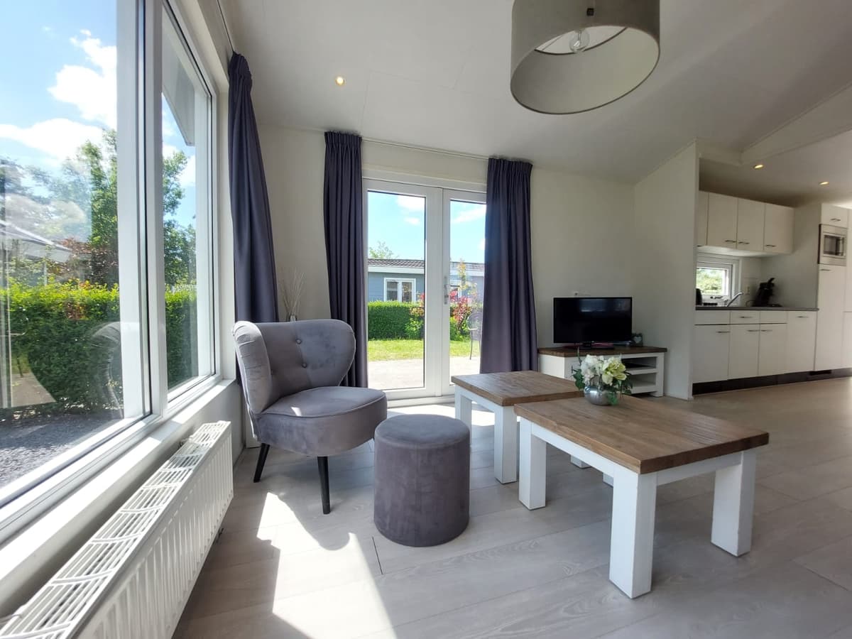 Moderne woonkamer met grote ramen, een stoel en een poef, houten tafel en open keuken in EuroParcs Bad Hulckesteijn.