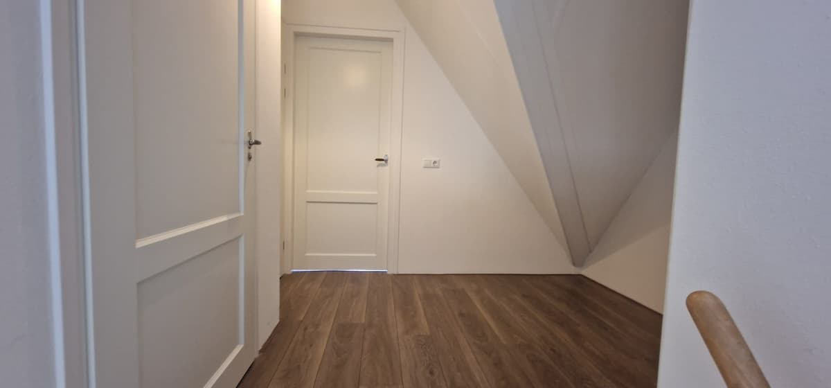 Lichte gang met een houten vloer, een deur naar een andere ruimte en schuine wanden.