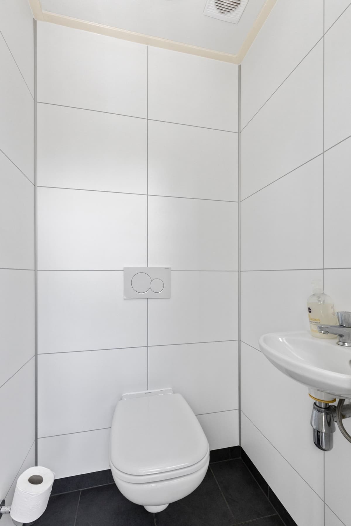 Witte toiletruimte met moderne tegels, een wastafel en een rol toiletpapier op de vloer.