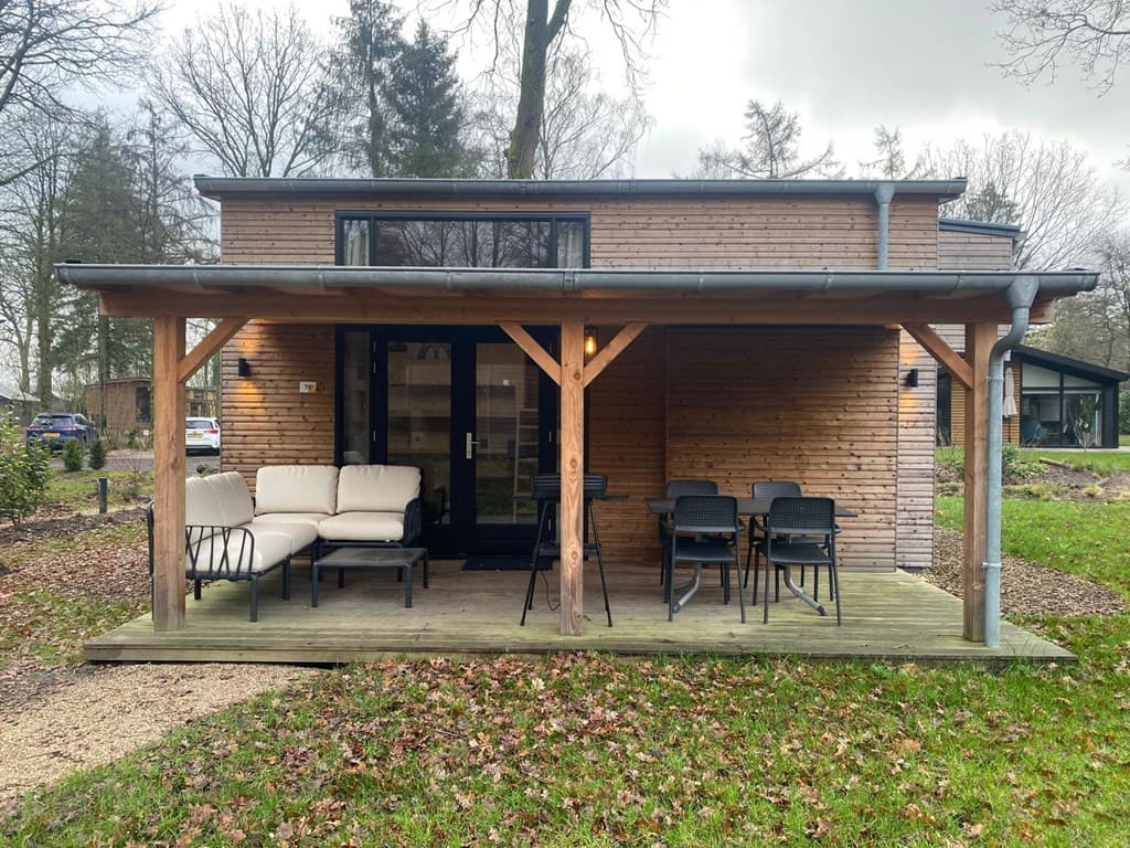 Moderne vakantiewoning met houten gevel en overdekt terras, omringd door bomen en gras.