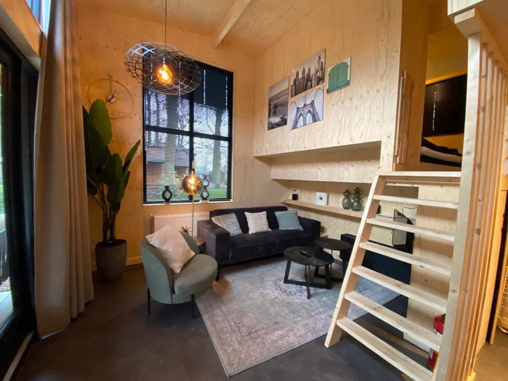 Knusse woonkamer met houten wand, bank en moderne verlichting, trap naar bovenverdieping en grote raampartij.