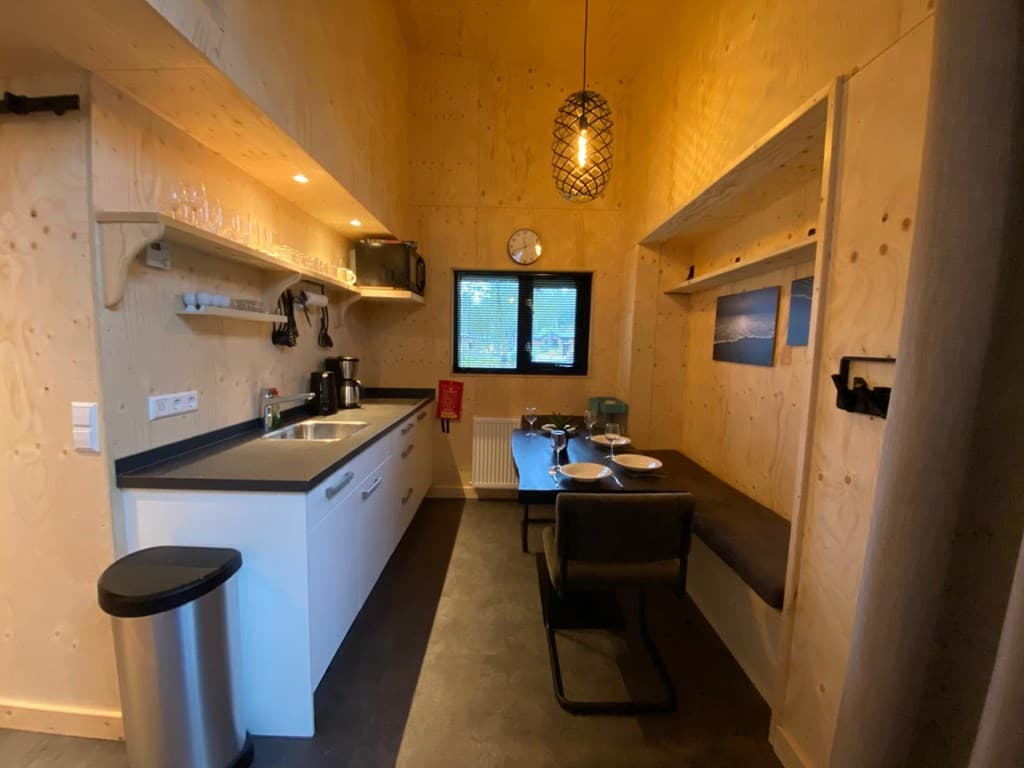 Keuken met houten wandafwerking, zwarte werkbladen, eettafel met stoelen en grote ramen voor natuurlijk licht