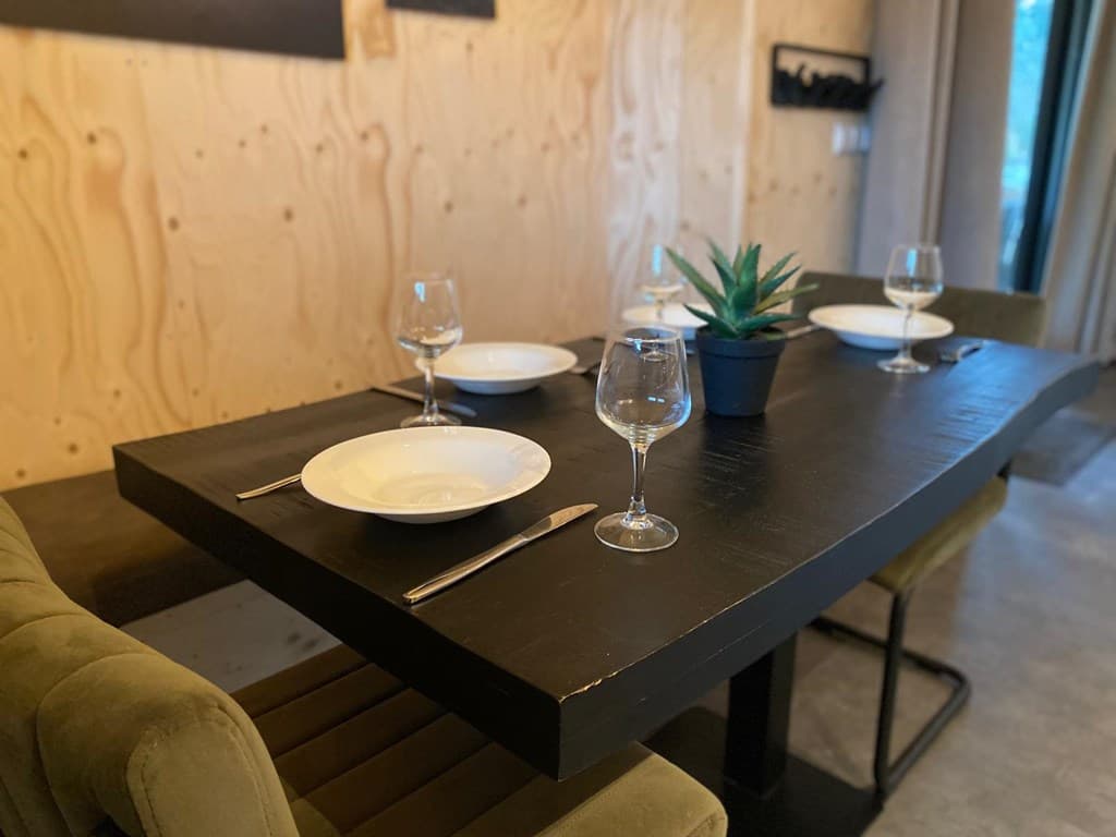 Eettafel met vier borden en glazen, omgeven door groene stoelen en een plant in pot, met houten wand op achtergrond