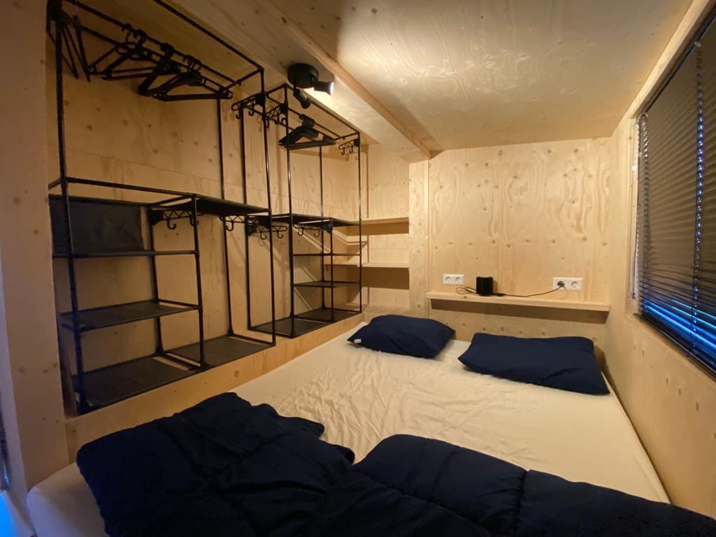 Slaapkamer met een tweepersoonsbed en open kledingrekken aan de wand van hout. Er zijn rolluiken voor het raam.