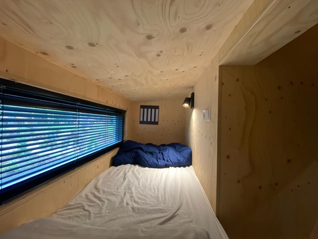 Slaapcabine met een bed, houten wanden en een raam met jaloezieën en verlichting aan de muur.