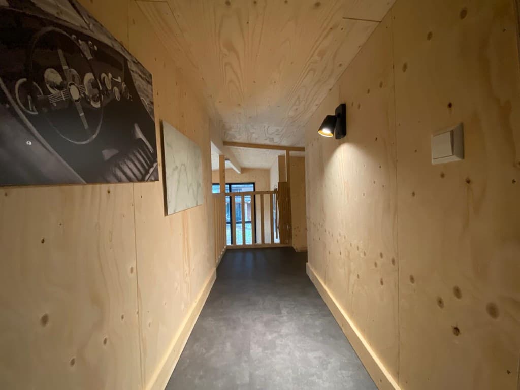 Corridor met houten wanden, verlichting aan het plafond en kunst aan de muren, leidt naar een open ruimte met ramen.
