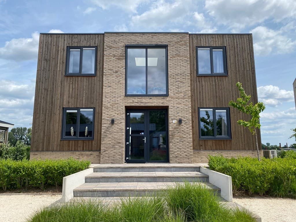 Moderne vakantiewoning met houten en stenen gevel, grote ramen en trap naar de ingang, omgeven door groen.