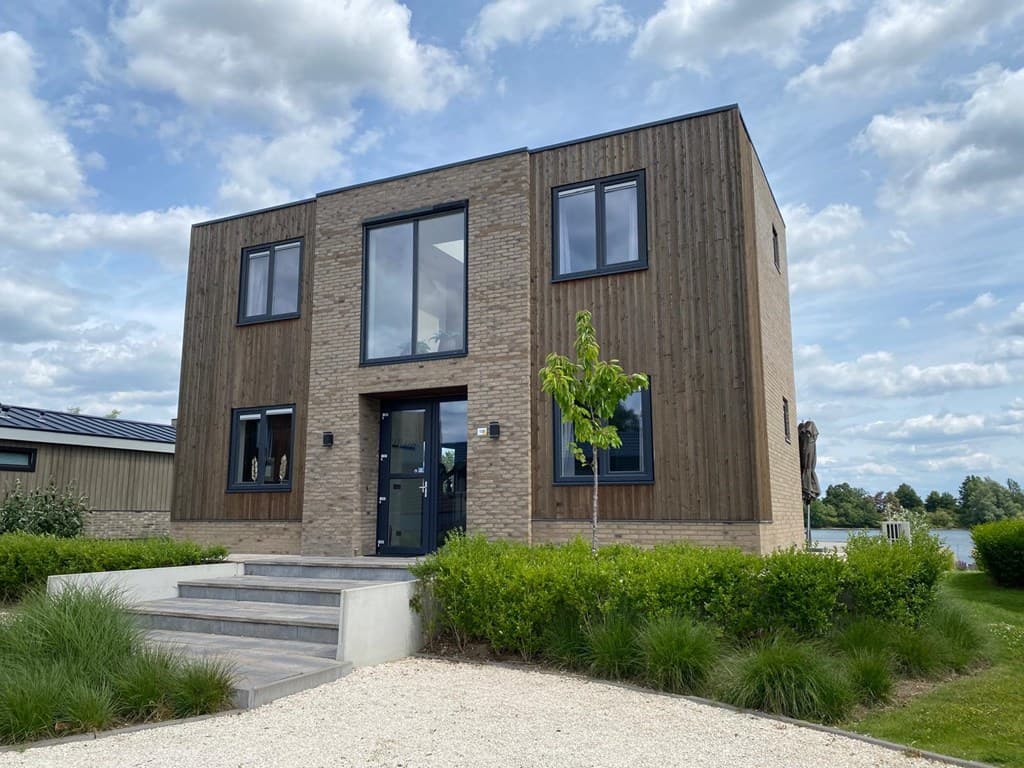 Moderne vakantiewoning met een combinatie van steen en hout, omgeven door groen en met een trap naar de voordeur.
