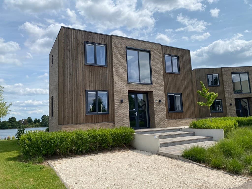 Moderne vakantiewoning met houten gevel en grote ramen, gelegen aan een grasveld bij het water.
