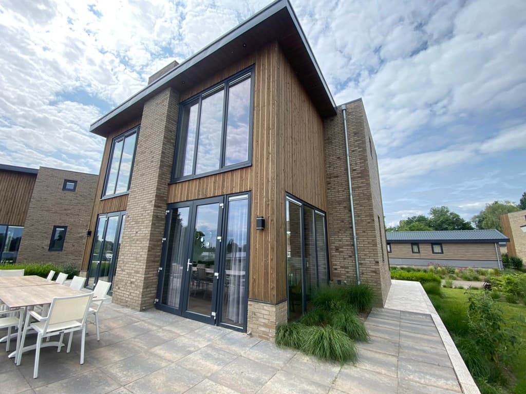 Moderne vakantiewoning met grote ramen, houten gevel en een betegeld terras met uitzicht op een groene tuin.
