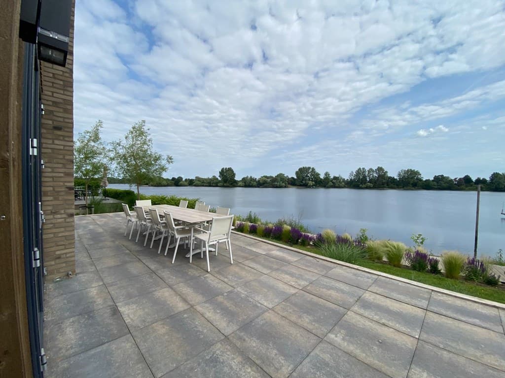 Zicht op terras met eettafel en stoelen aan de Maas, omgeven door groen en een heldere lucht met wolken.