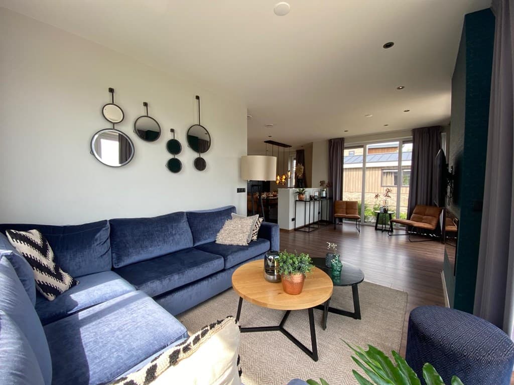 Moderne woonkamer met blauwe bank, ronde spiegels en een houten tafel, met zicht op een keuken en raam met veel licht.