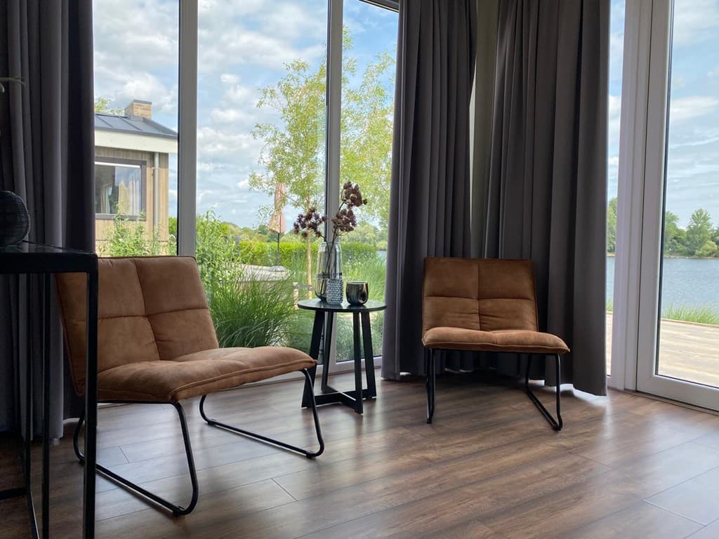 Twee comfortabele zitstoelen met uitzicht op water en natuur door grote ramen met gordijnen.