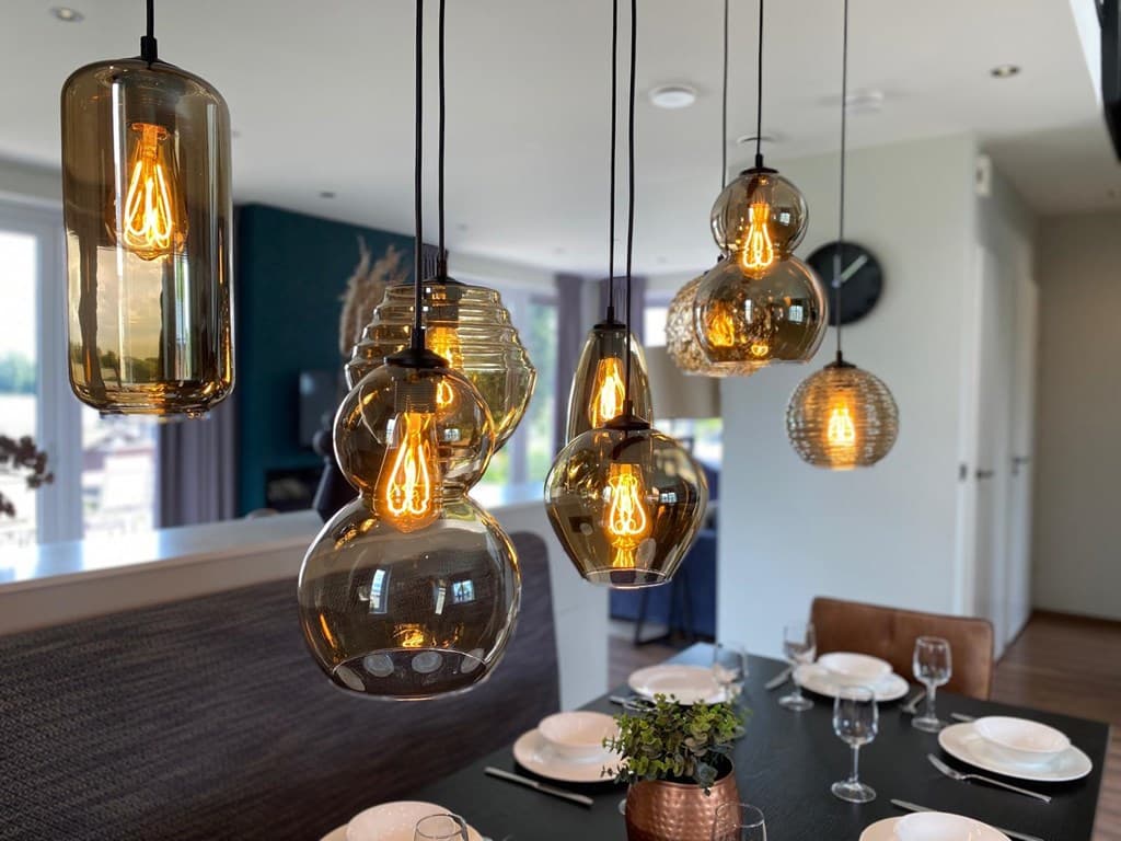 Designlampen hangen boven een gedekte eettafel met glazen en borden, lichte muren en grote ramen op de achtergrond.