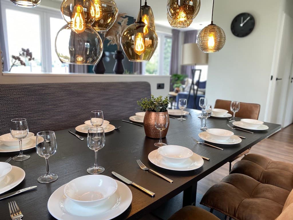 Eettafel met 12 borden en glazen, sfeervolle hanglampen en een decoratieve plant in een vakantiehuis aan de Maas.