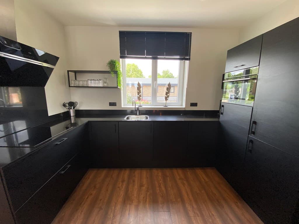 Moderne zwarte keuken met een raam, spoelbak, en inbouwapparatuur op een houten vloer.