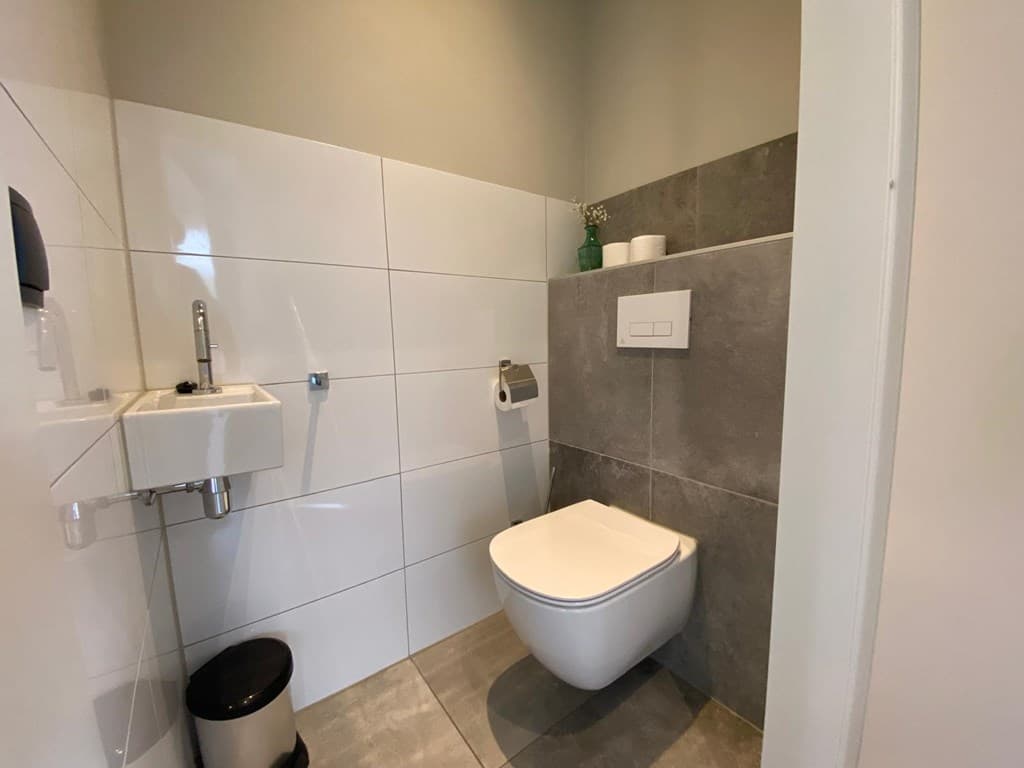 Moderne toiletruimte met witte en grijze wandtegels, een staand toilet en een klein fonteintje aan de muur.
