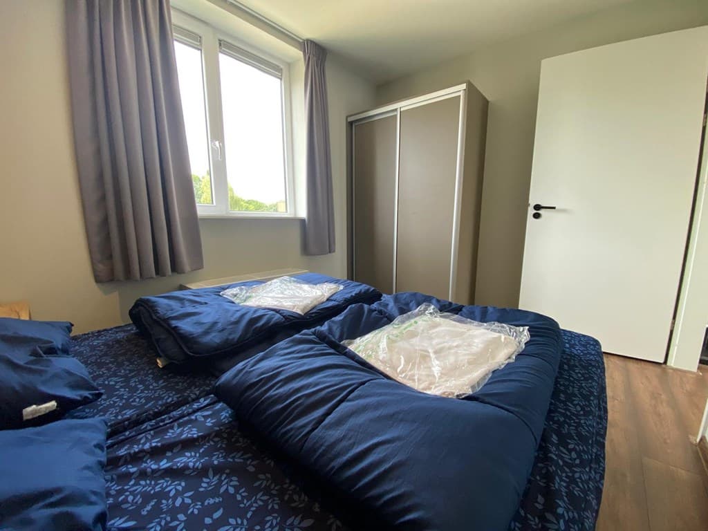 Slaapkamer met een tweepersoonsbed en een kledingkast, lichtinval vanuit een raam en grijze gordijnen.