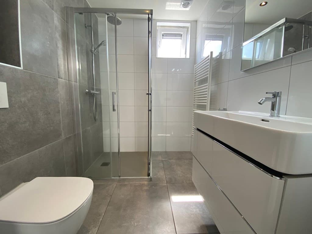 Moderne badkamer met inloopdouche, wastafel met kast en toilet tegen een grijze wand.