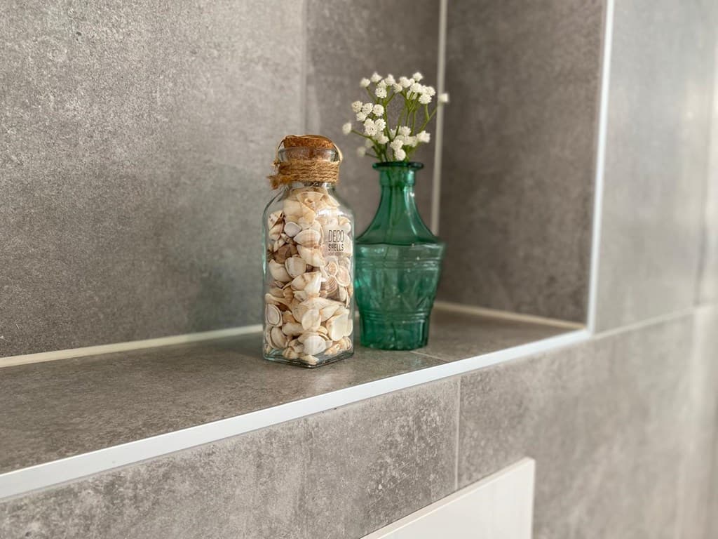 Decoratieve glazen fles met schelpen en een groene vaas met bloemen op een grijze tegelvloer