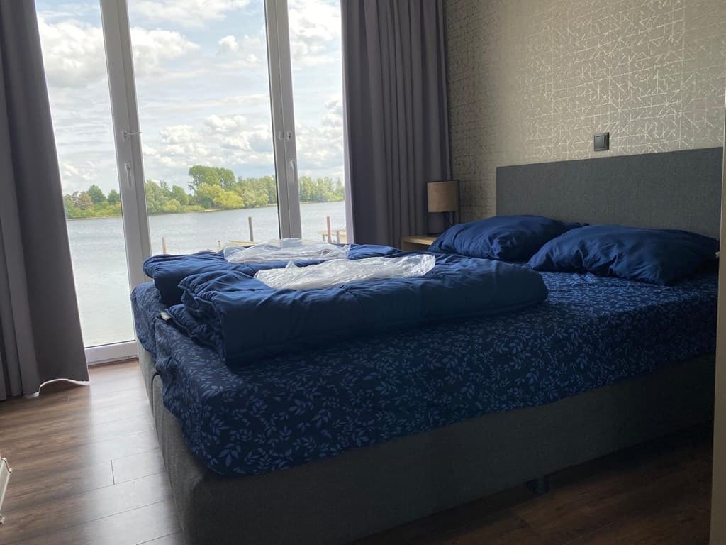 Slaapkamer met groot bed en uitzicht op het water door glazen deur en raam, gordijnen omhoog.
