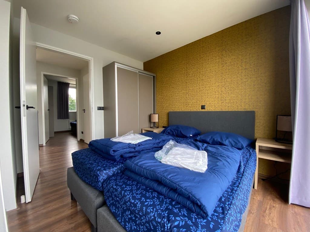 Slaapkamer met een grijze bed met blauwe dekbedden, een nachttafeltje en een kast met een gele wand achter het bed