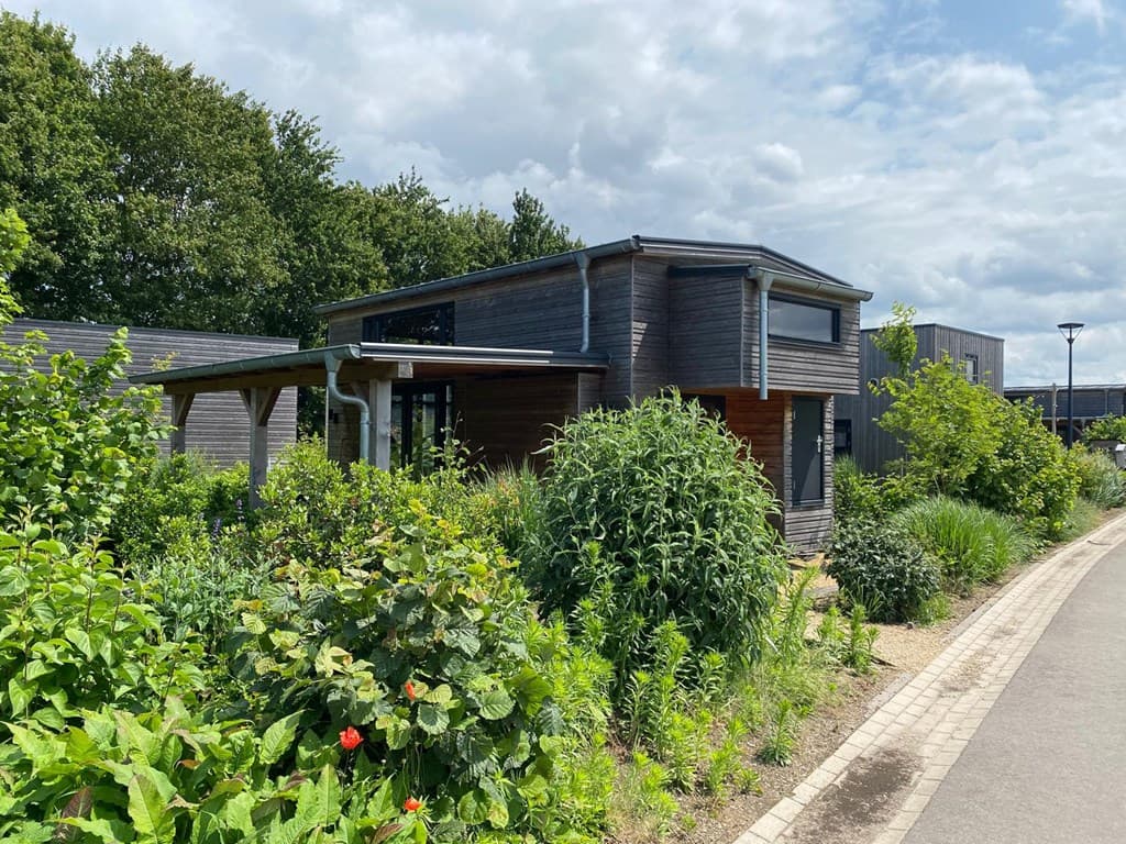 Moderne vakantiewoning omgeven door groen en planten, met een houten gevel en een overdekte ingang.