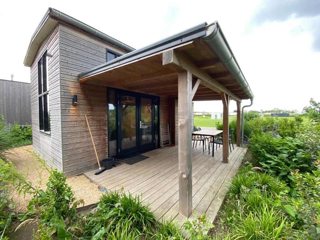 Houten vakantiehuis met overdekt terras en tuin, gelegen in een rustige omgeving met groene struiken rondom.