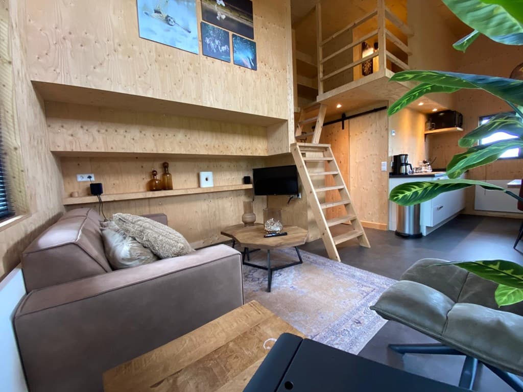 Interieur van een vakantiewoning met houten wanden, zithoek, tv, ladder naar de slaapverdieping en planten.