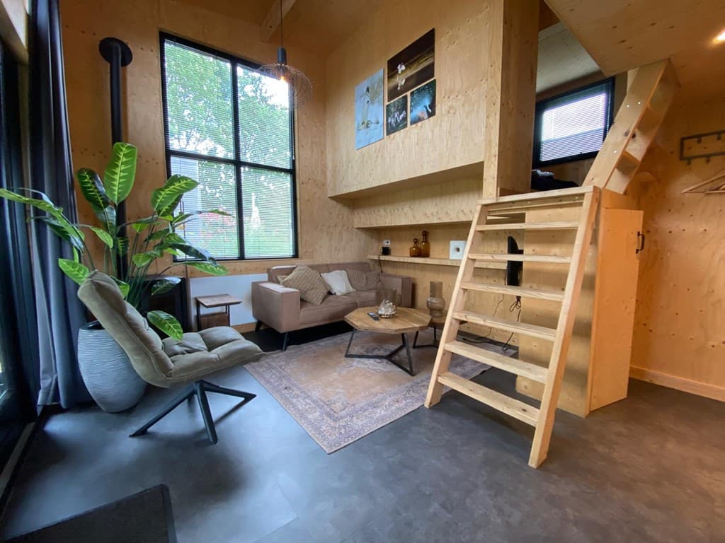Interieur van een vakantiewoning met hoge ramen, een zithoek met bank en fauteuil, en een houten trap naar een bovenverdiepin