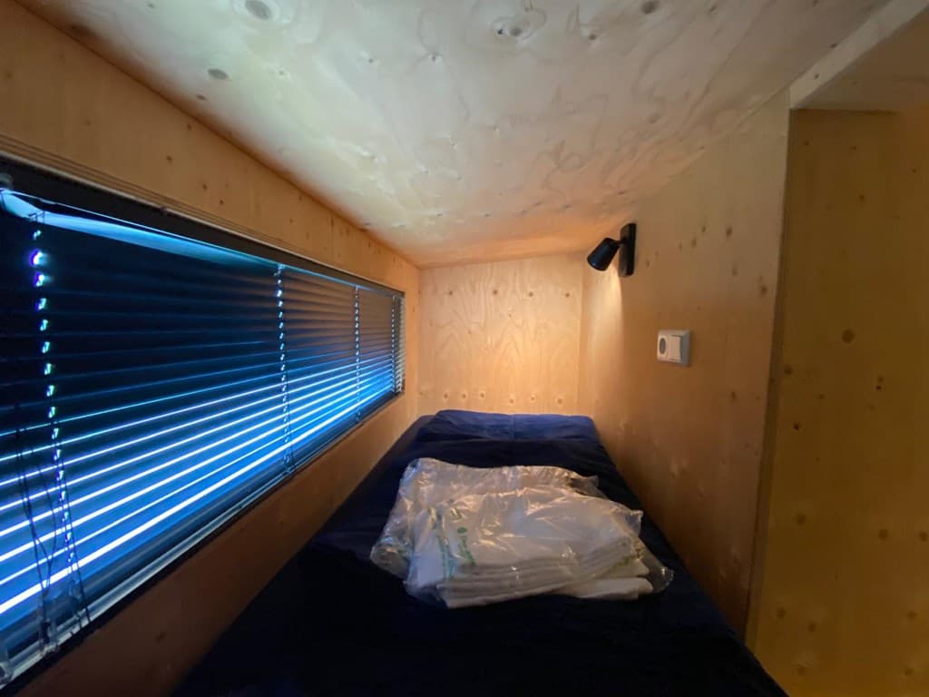 Slaapruimte met houten wanden, een enkel bed, een raam met jaloezieën en gestapeld beddengoed op de matras