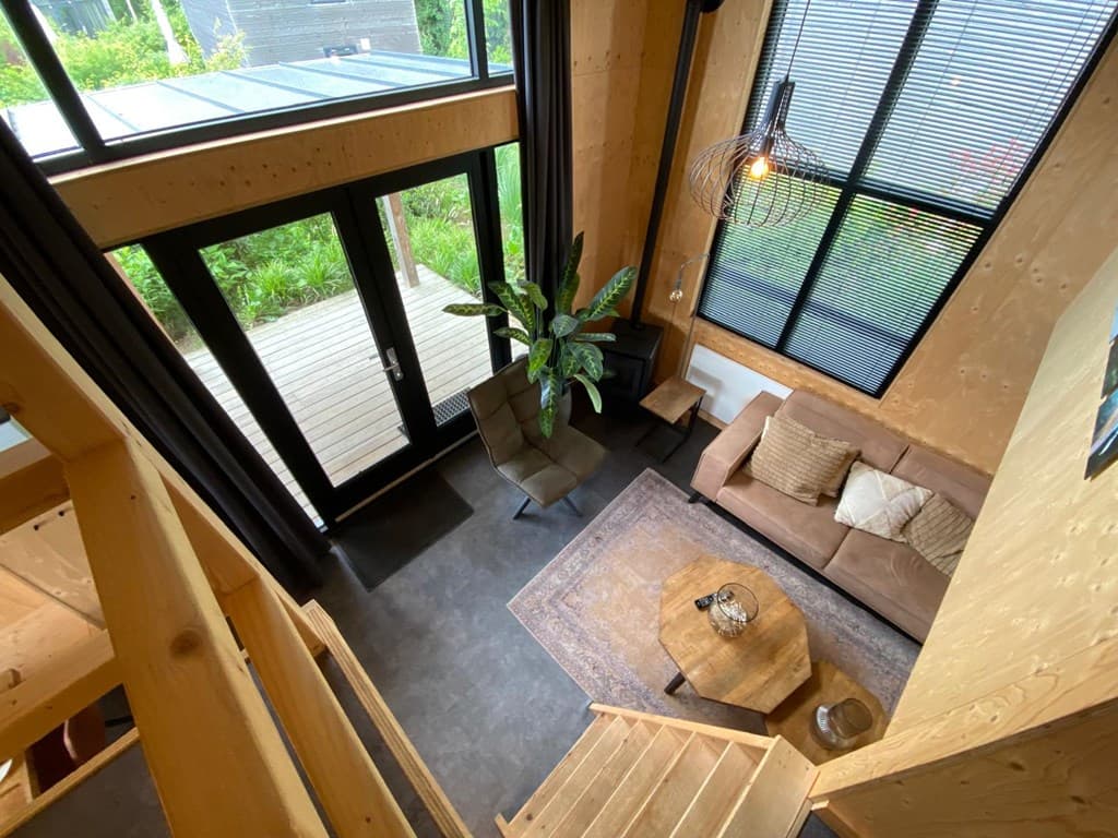 Ruime zithoek met bank, tafel en planten, grote ramen en toegang tot een terras met houten vloer.