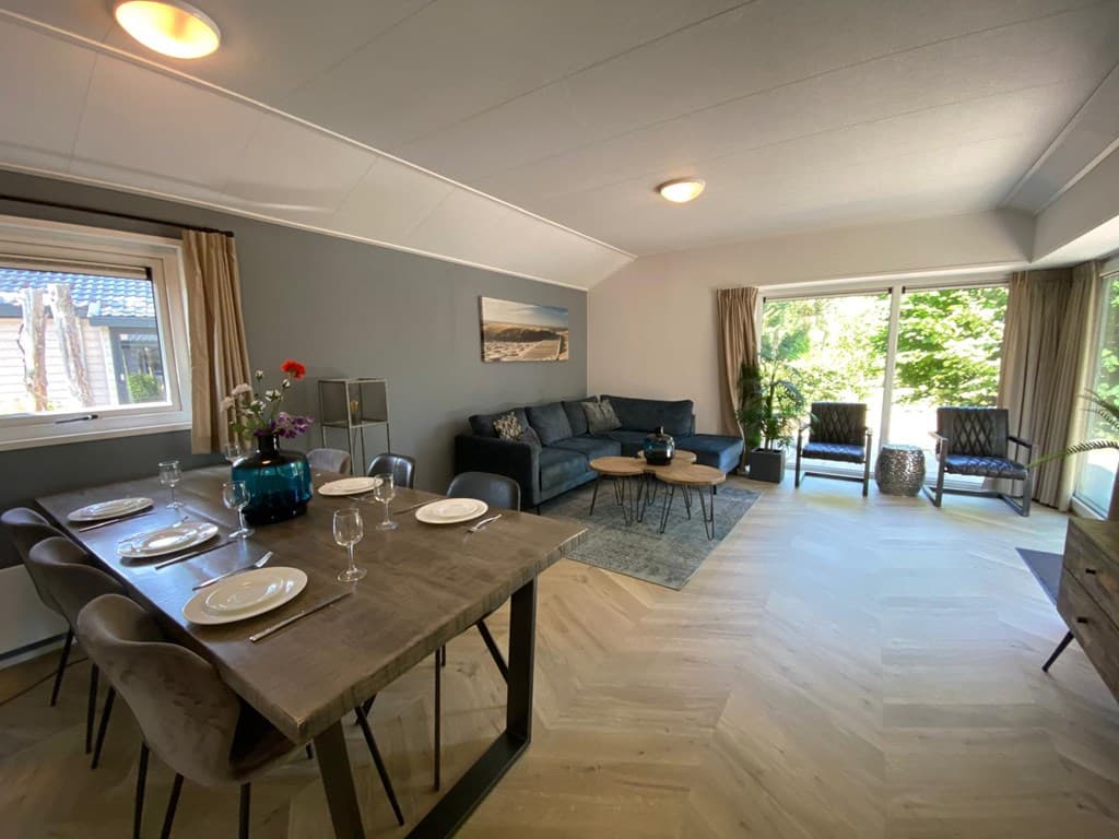 Zithoek met banken en fauteuils, naast een eettafel voor zes personen en grote ramen met uitzicht op de groene omgeving.