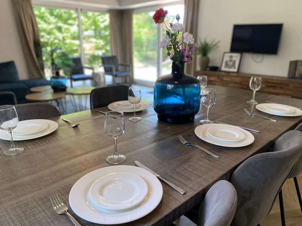Eetkamer met een grote houten tafel, gedekte borden, glazen en een blauwe vaas met bloemen op de tafel