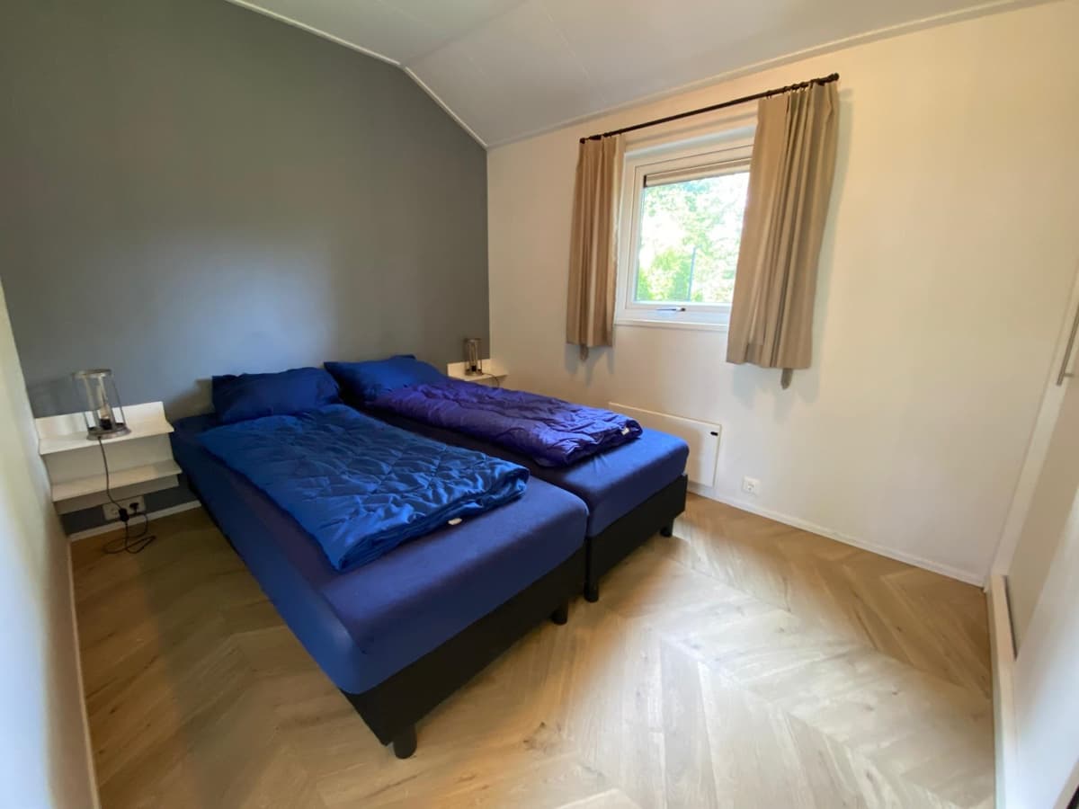 Bedkamer met twee losse bedden, blauwe dekbedden en raam met gordijnen. Houten vloer en neutrale wandkleur.